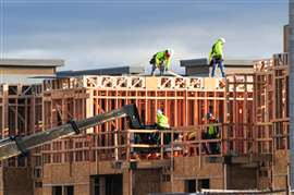 Construction workers frame a multi-unit residential building, working on scaffolding and rooftops as part of an active housing development project in East Mesa, Arizona