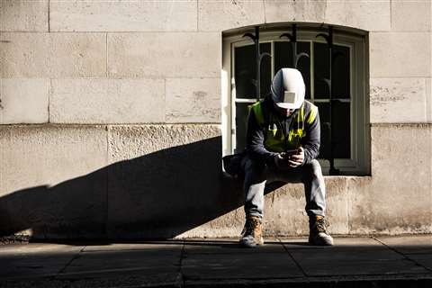 A lone construction worker hearing PPE stares at his phone while sitting on a stone window sill